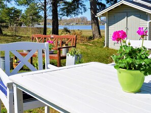 Et hvidt bord med en grøn potteplante, en træstol og et skur i en græsmark med træer og et vandareal i baggrunden.
