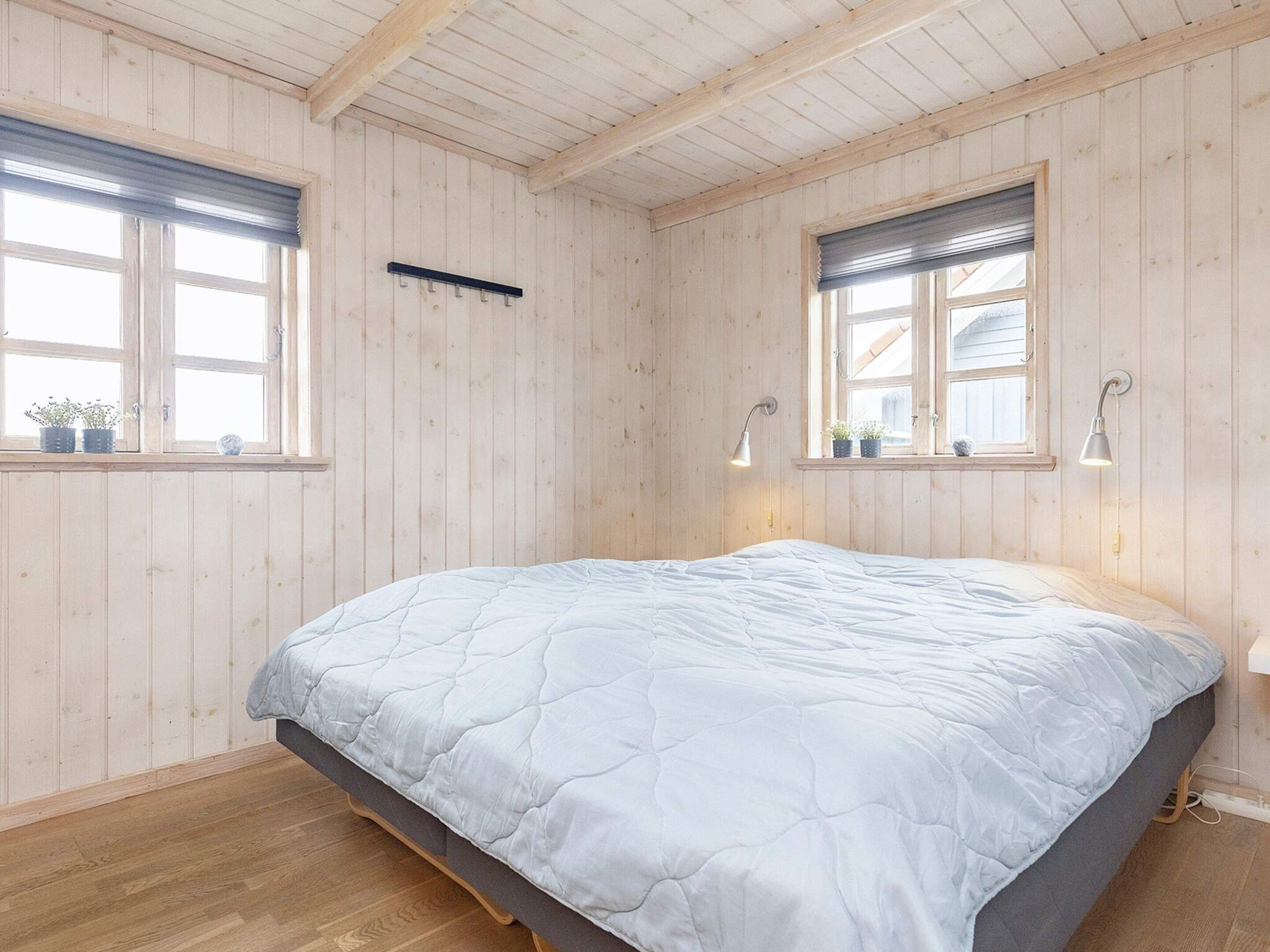 A bedroom with a bed, wooden walls, two windows with blinds, and bedside lamps.