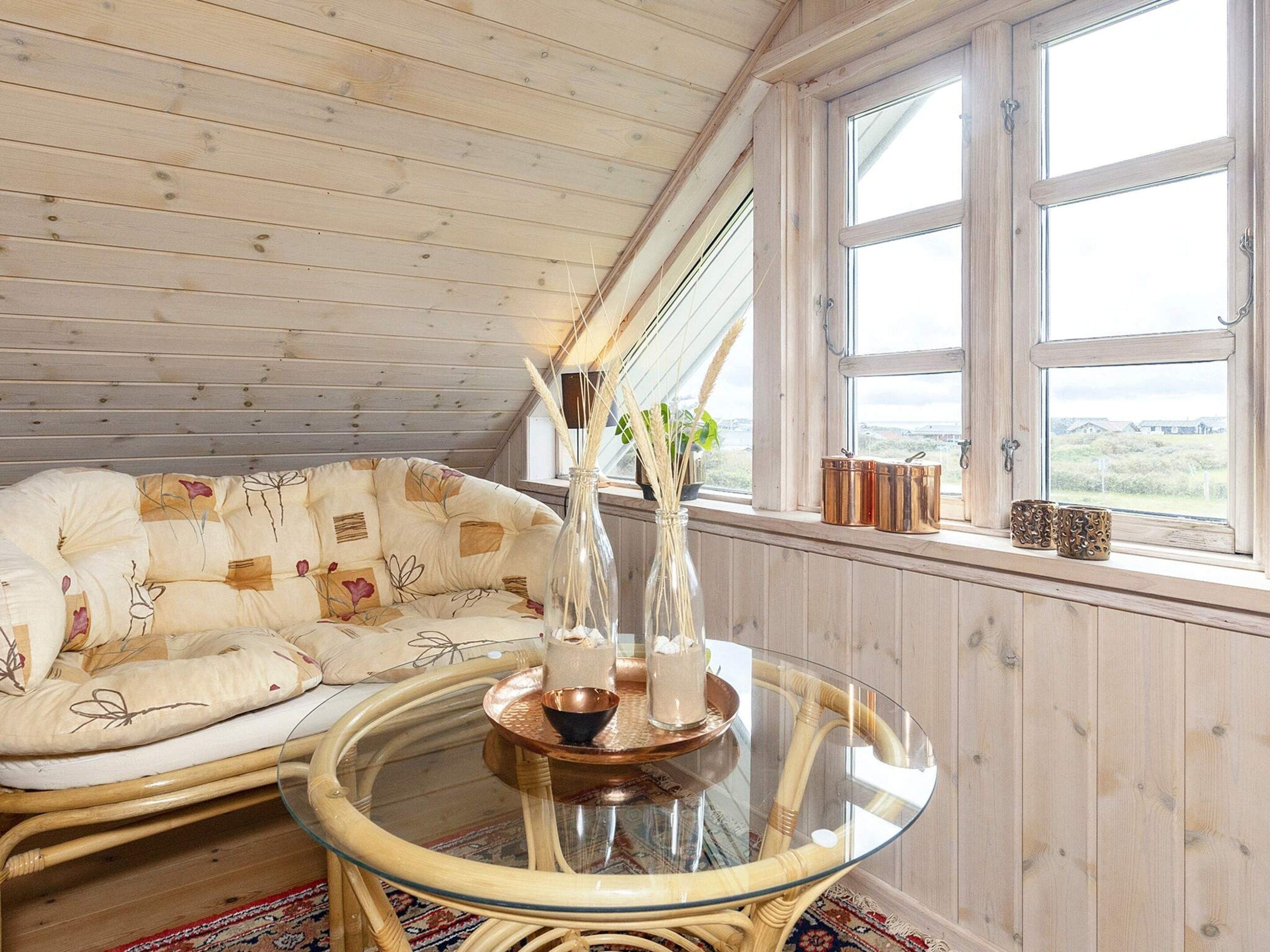 A cozy room with a wooden ceiling, a cushioned sofa, a glass table with a vase, and a window with a view.