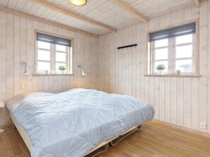 Une pièce aux murs en bois, un lit recouvert d’une couette blanche, deux fenêtres équipées de stores et un luminaire suspendu.