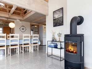 A modern interior with a dining area featuring a wooden table and chairs, a black stove with a visible fire, and a wall clock.