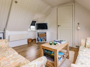 A cozy room with a sloped ceiling, a television on a white stand, a bookshelf, a wooden table with puzzle games, and a door.