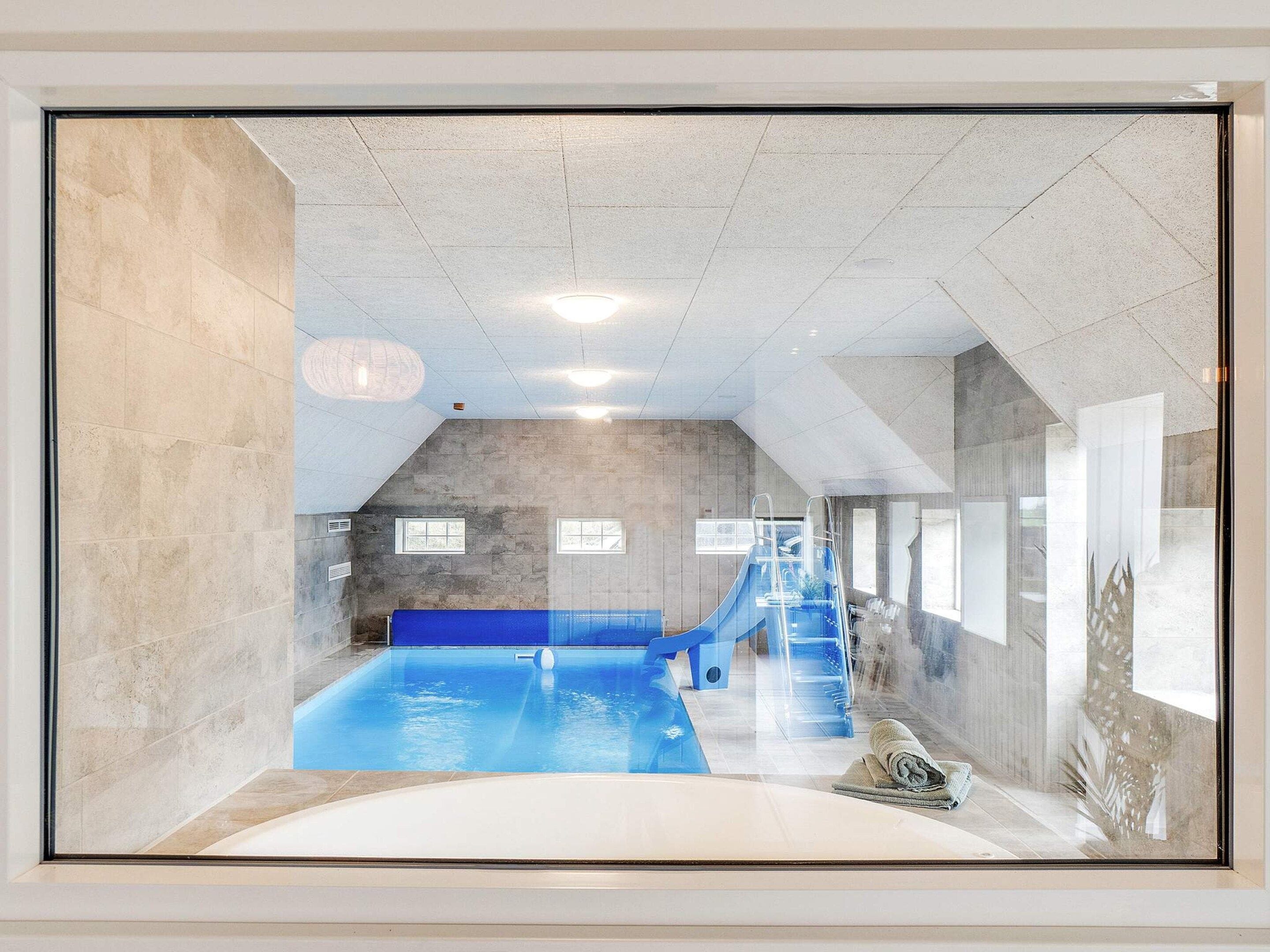 Une piscine intérieure dotée d’un toboggan bleu, d’un plongeoir et d’un espace salon aménagé en contrebas.