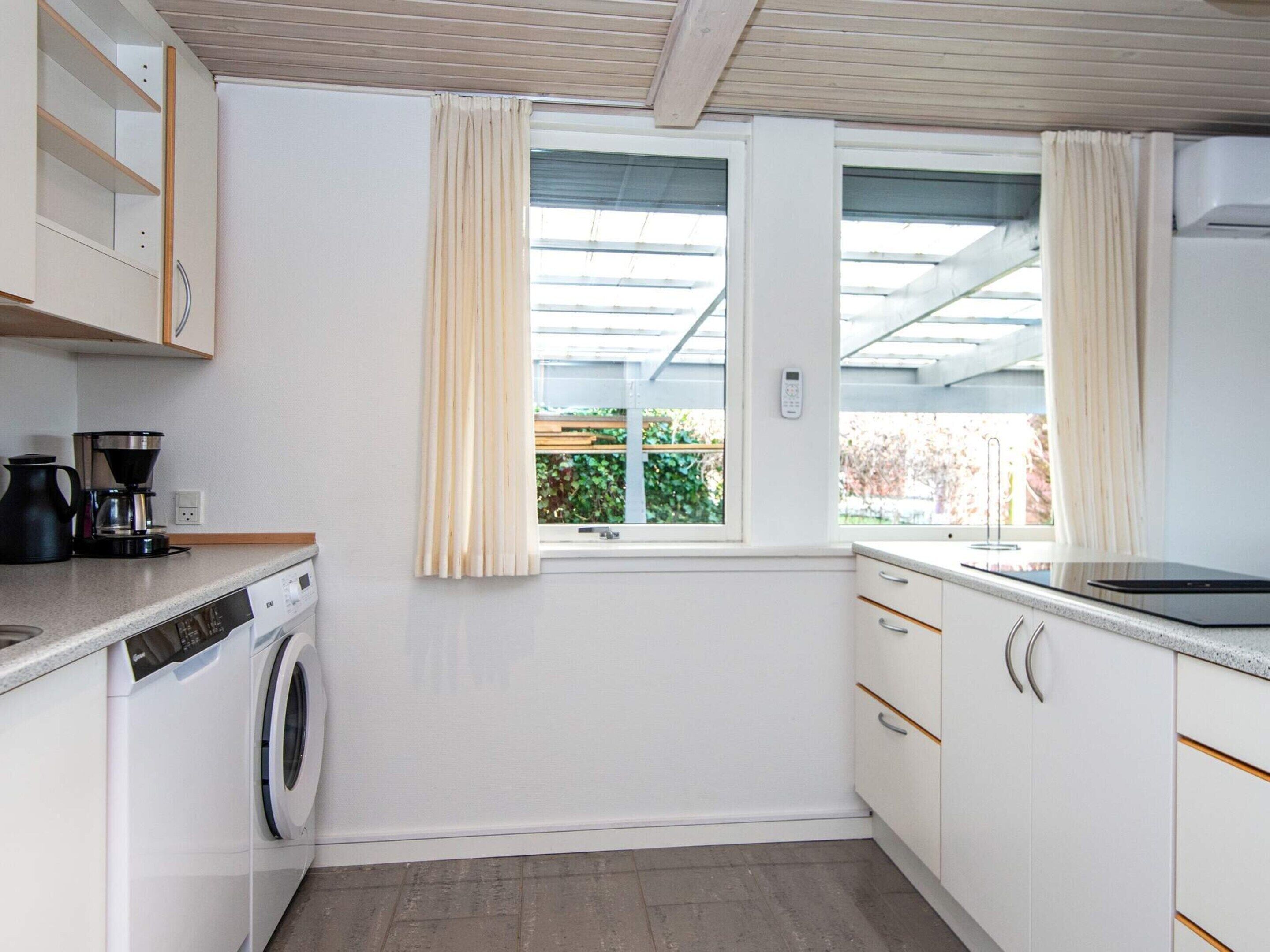 Cocina compacta con lavadora, cafetera y ventana con cortinas.