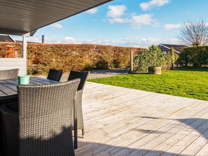 Um alpendre coberto com deck de madeira, móveis de vime e vista para um jardim e gramado bem cuidados.