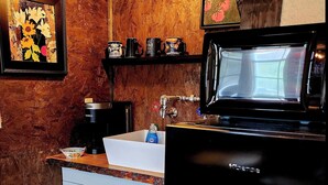 A compact kitchen with a white sink, a coffee maker, and a microwave. There are framed artworks on the walls and a shelf with mugs. - The Artists Cottage ~ Fun desert getaway (Ajo)
