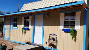 A small, single-story building with a beige exterior and blue trim. It features a front door, two windows with shutters, and a small porch area. - The Artists Cottage ~ Fun desert getaway (Ajo)