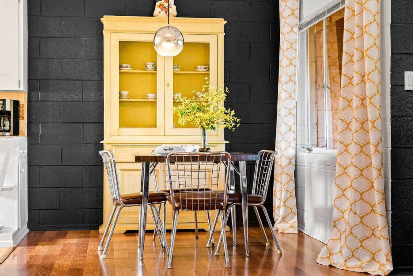 A dining area with a table and chairs, a yellow cabinet with glass doors, and a window with patterned curtains.