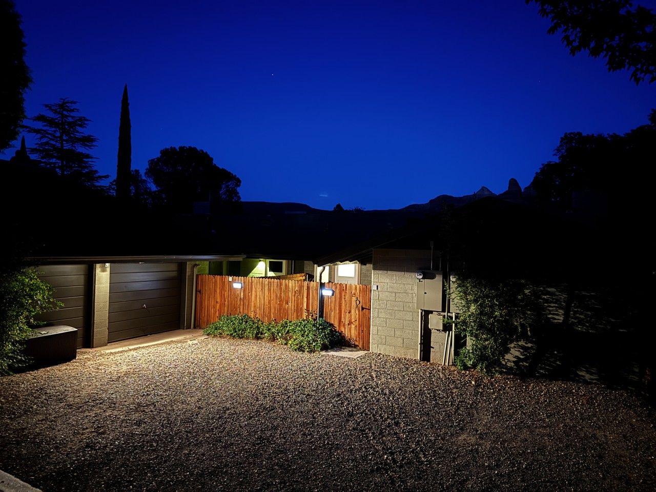 Un garage attenant à une maison, avec une allée en gravier, une clôture en bois et un grand arbre en arrière-plan.