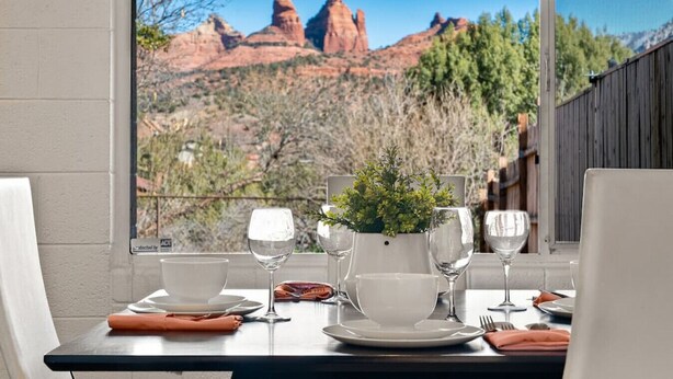 Mesa servida con platos, tazones y copas de vino, con vista a formaciones rocosas rojas a trav és de una ventana.