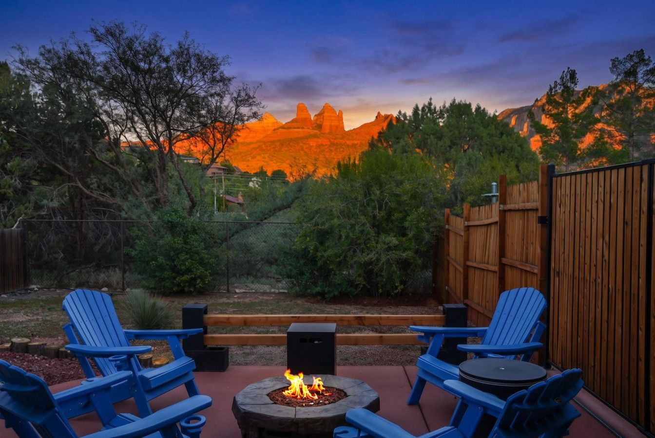 An outdoor seating area with Adirondack chairs and a fire pit, surrounded by a wooden fence and vegetation.