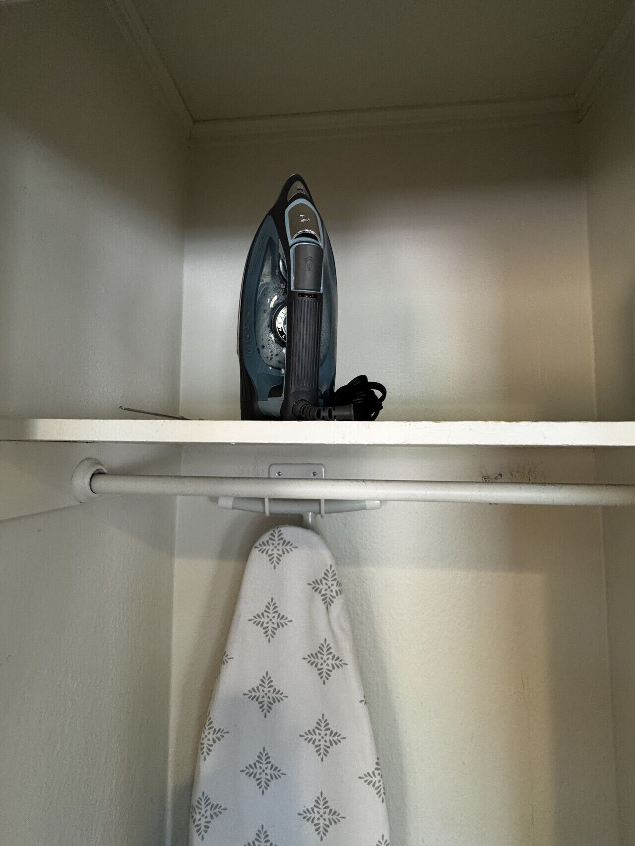 A storage closet with a hanging rack and an iron on the top shelf.