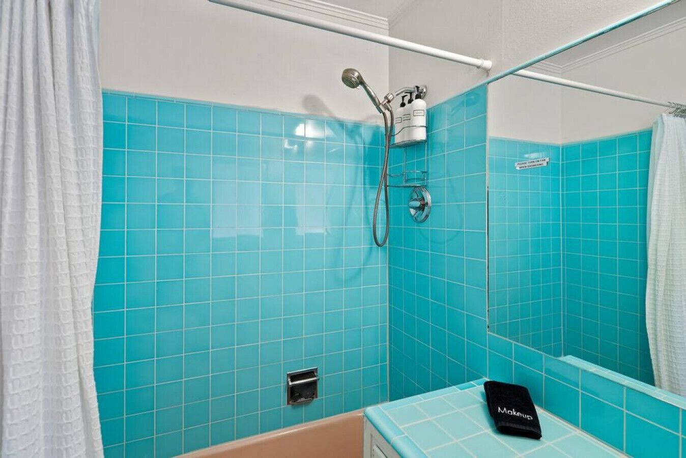 Une salle de bain avec des carreaux turquoise, une douche équipée d’un pommeau de douche amovible, un porte-serviettes et un lavabo avec un distributeur de savon.
