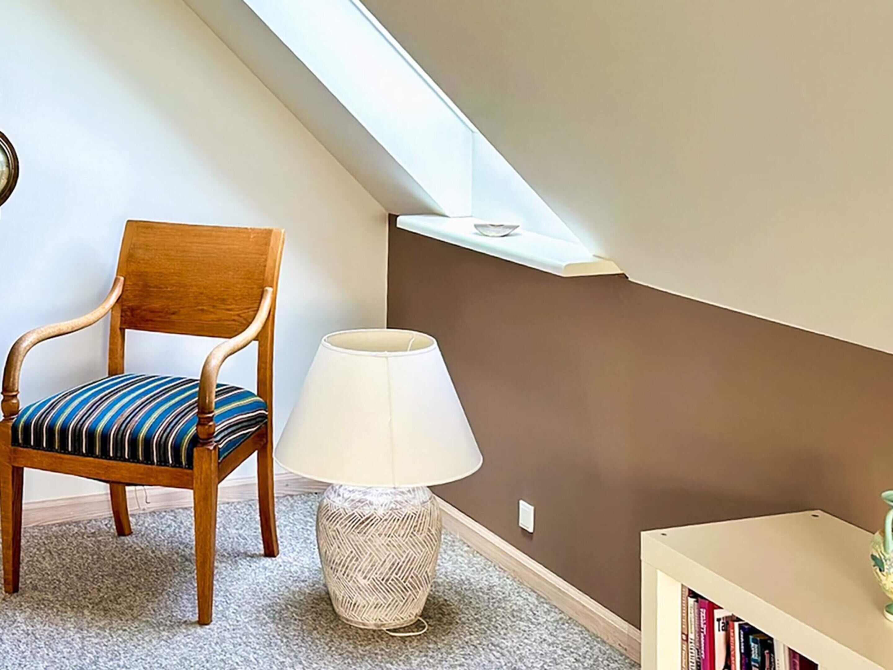 A room with a wooden chair, a woven floor lamp, a bookshelf, and a slanted ceiling with a skylight.