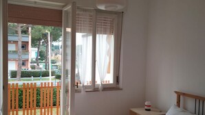 A bedroom with a bed, a bedside table, a window with curtains, and a view of a balcony with a fence. -  M335 - Marcelli, large two-room apartment with sea view with balcony (Marcelli)