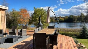 An outdoor deck with a wooden table and chairs, a hanging lamp, and a view of a lake surrounded by trees.