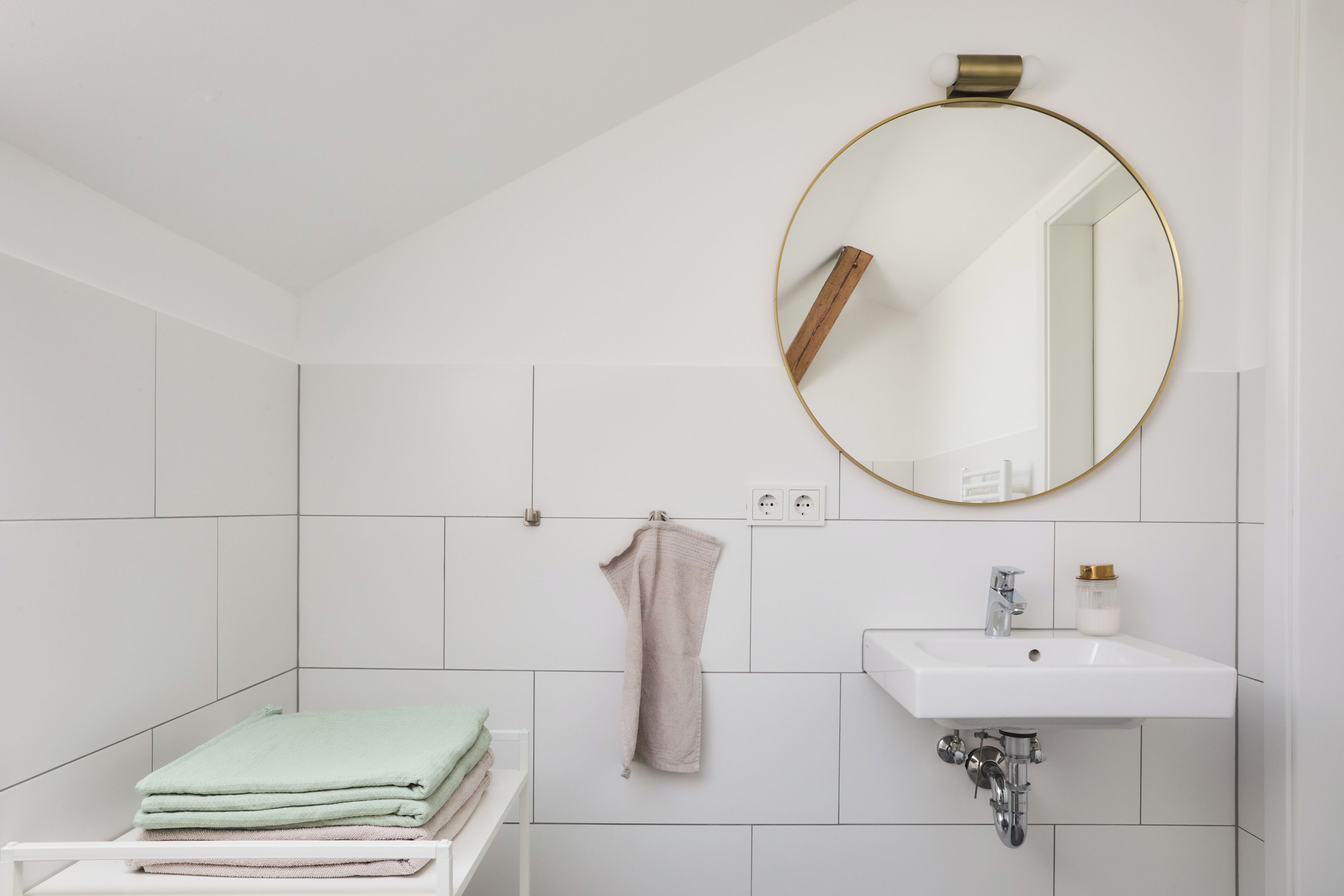 Un baño con baldosas blancas, un espejo redondo con marco dorado, un lavabo y toallas ordenadas en una estantería.