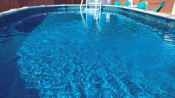 Une piscine aux eaux claires, avec un escalier permettant d’y accéder, entourée d’une clôture en bois et de quelques chaises vertes.
