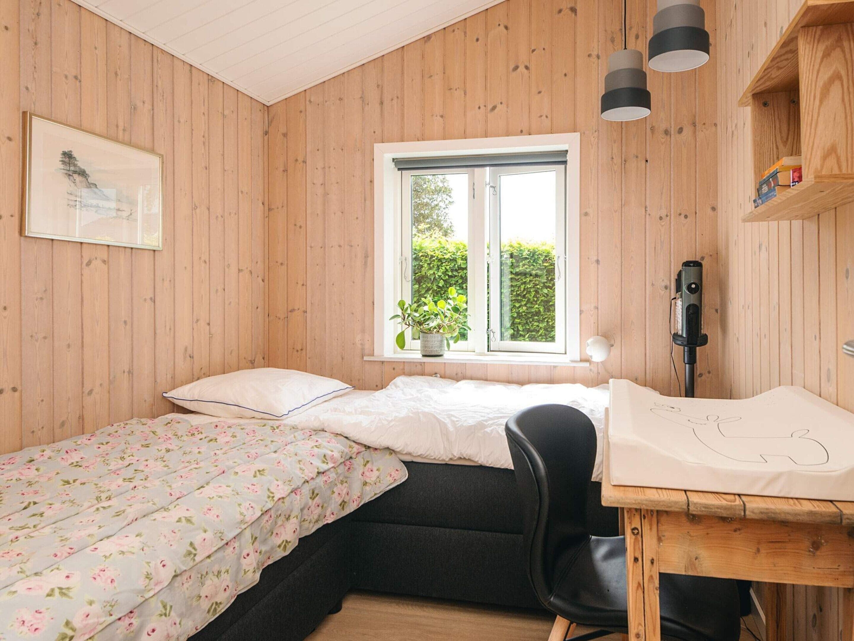 A room with wooden walls, a bed with a floral bedspread, a desk with a chair, a window with a plant, and a mounted device.