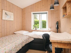 Ein Zimmer mit Holzwänden, einem Bett mit floralem Überwurf, einem Schreibtisch mit Stuhl, einem Fenster mit Pflanze und einem angebrachten Gerät.