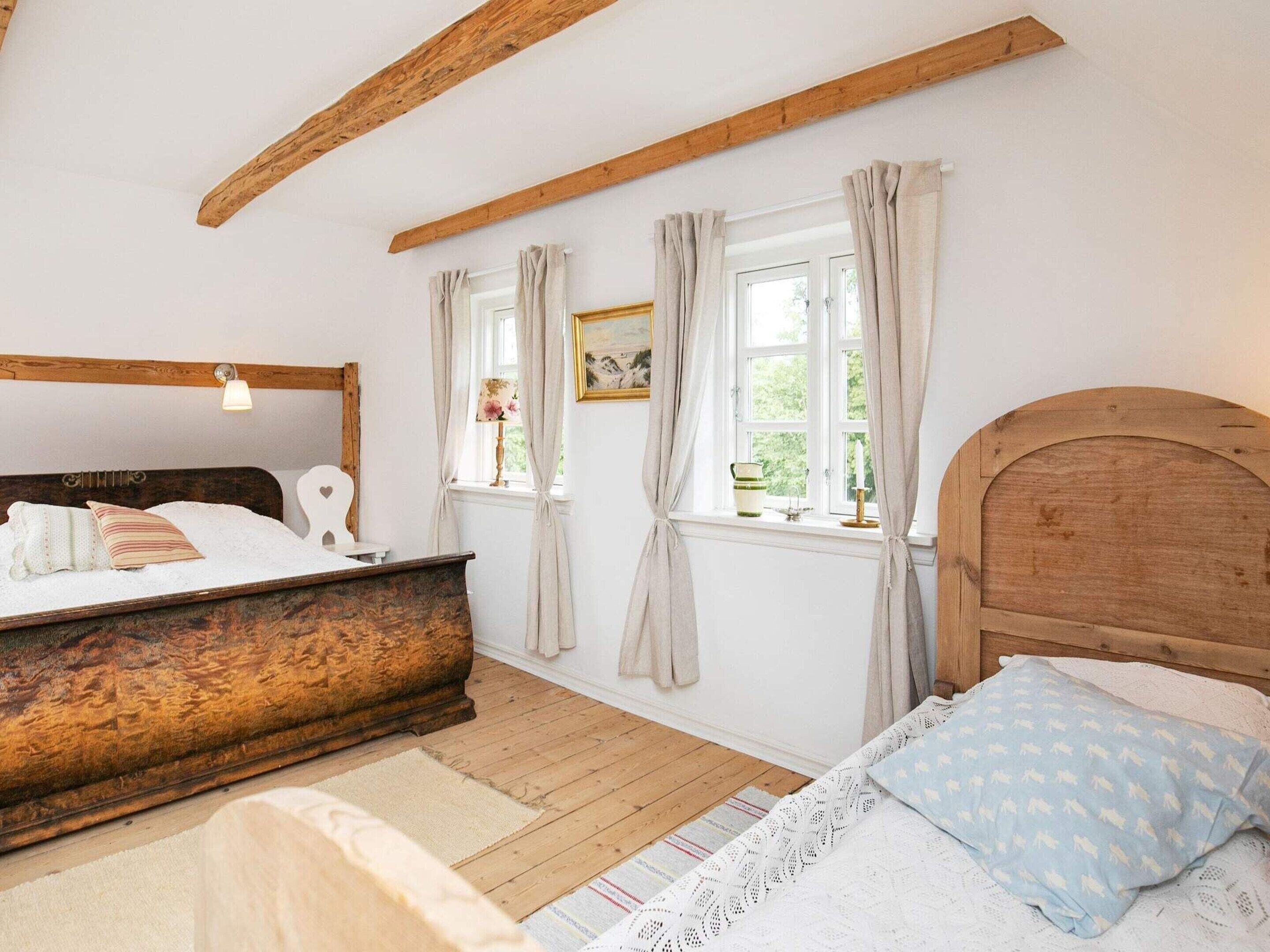 A bedroom with a wooden bed frame, a bedside table, a window with curtains, and a view of greenery outside.