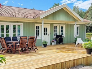 A green house with a covered patio, a barbecue grill, and a dining area with wooden chairs and a table. - 4 Star Holiday Home in Ellos (Ellos)