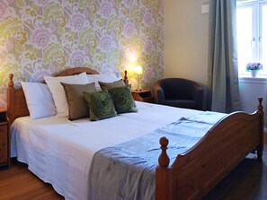 A neatly made bed with a wooden headboard and footboard, a bedside table with a lamp, and a chair in a room with patterned wallpaper. - 4 Star Holiday Home in Ellos (Ellos)