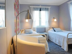 A room with two beds, a bedside table, a lamp, and a window with curtains.
