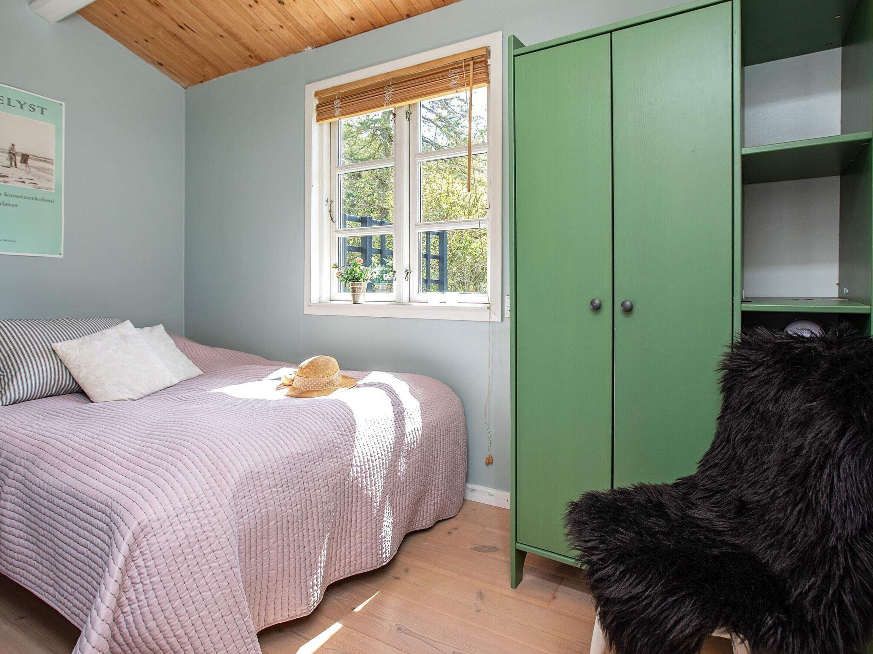 A bedroom with a bed, a green wardrobe, a window with blinds, and a hat on the bed.
