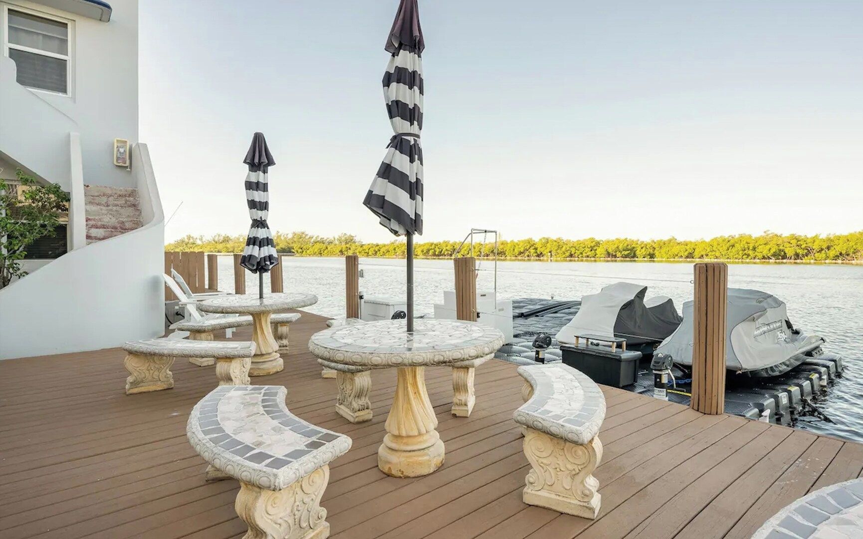Une terrasse avec des tables et des chaises en pierre, un bateau, et une vue sur une étendue d’eau bordée de verdure.