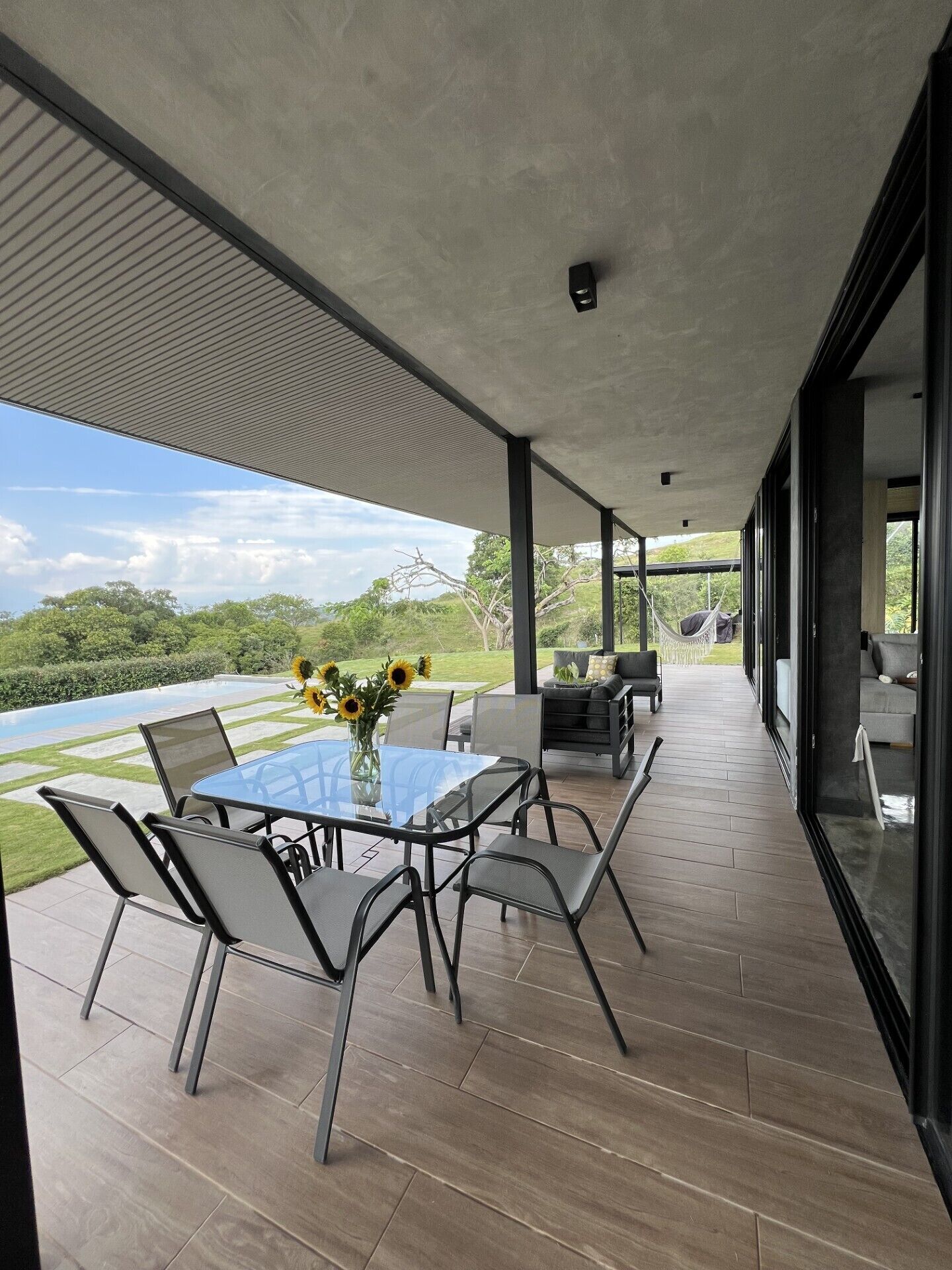 Un espacio moderno para comer al aire libre, con una mesa de vidrio y sillas de metal, con vistas a un jardín ajardinado y una masa de agua.