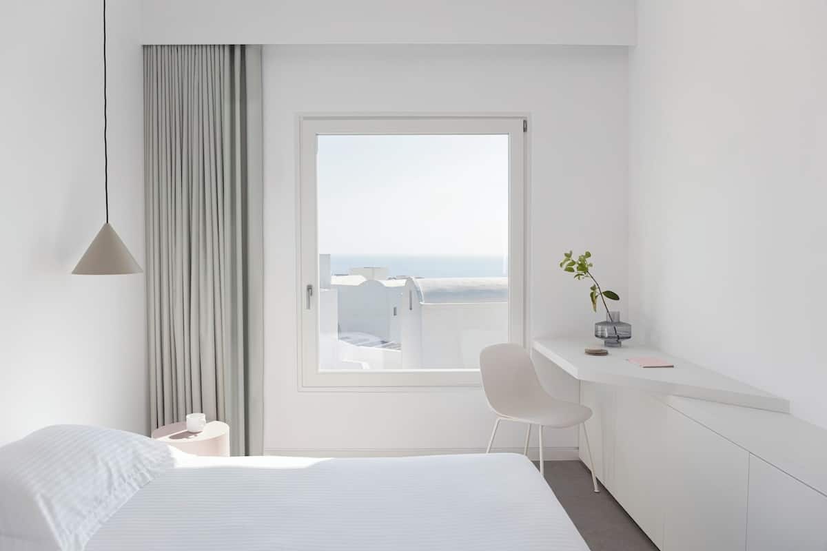 A minimalist hotel room with a large bed, a desk with a chair, a potted plant, and a view of the sea through a large window.