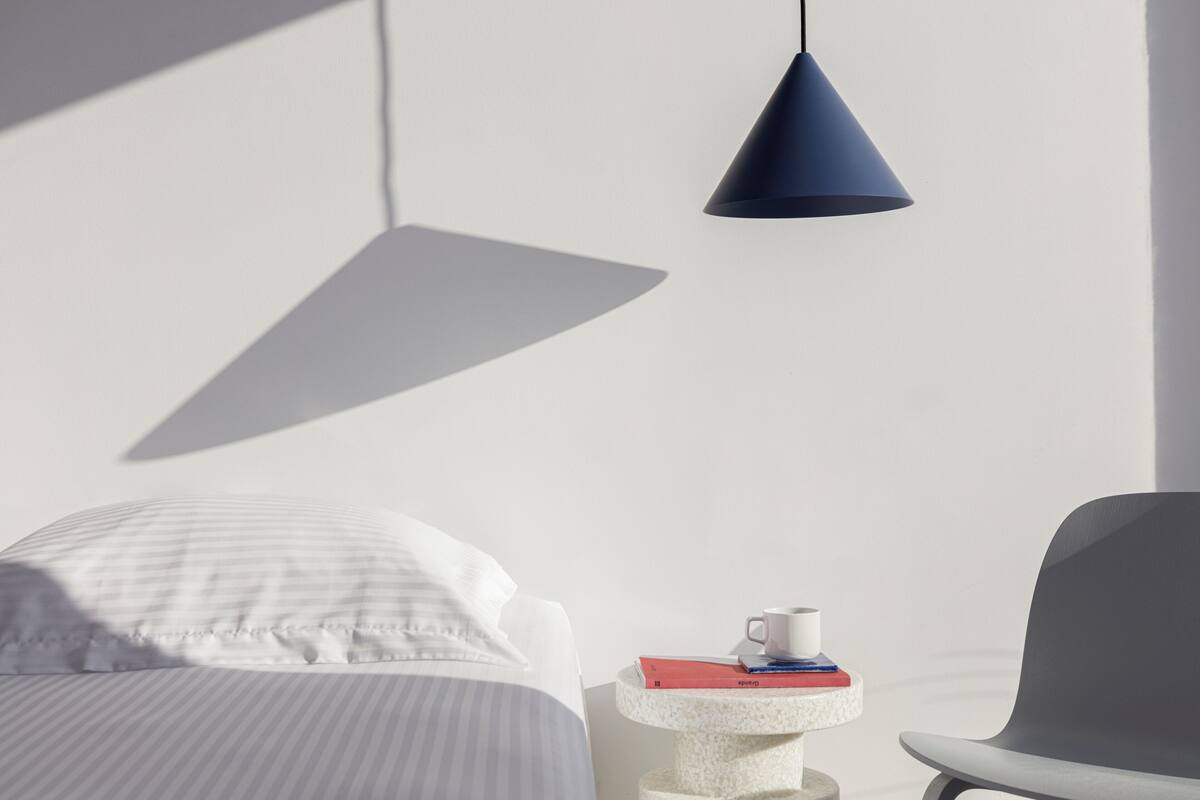 A modern bedroom with a bed, a bedside table with a cup and books, a chair, and a hanging light fixture.