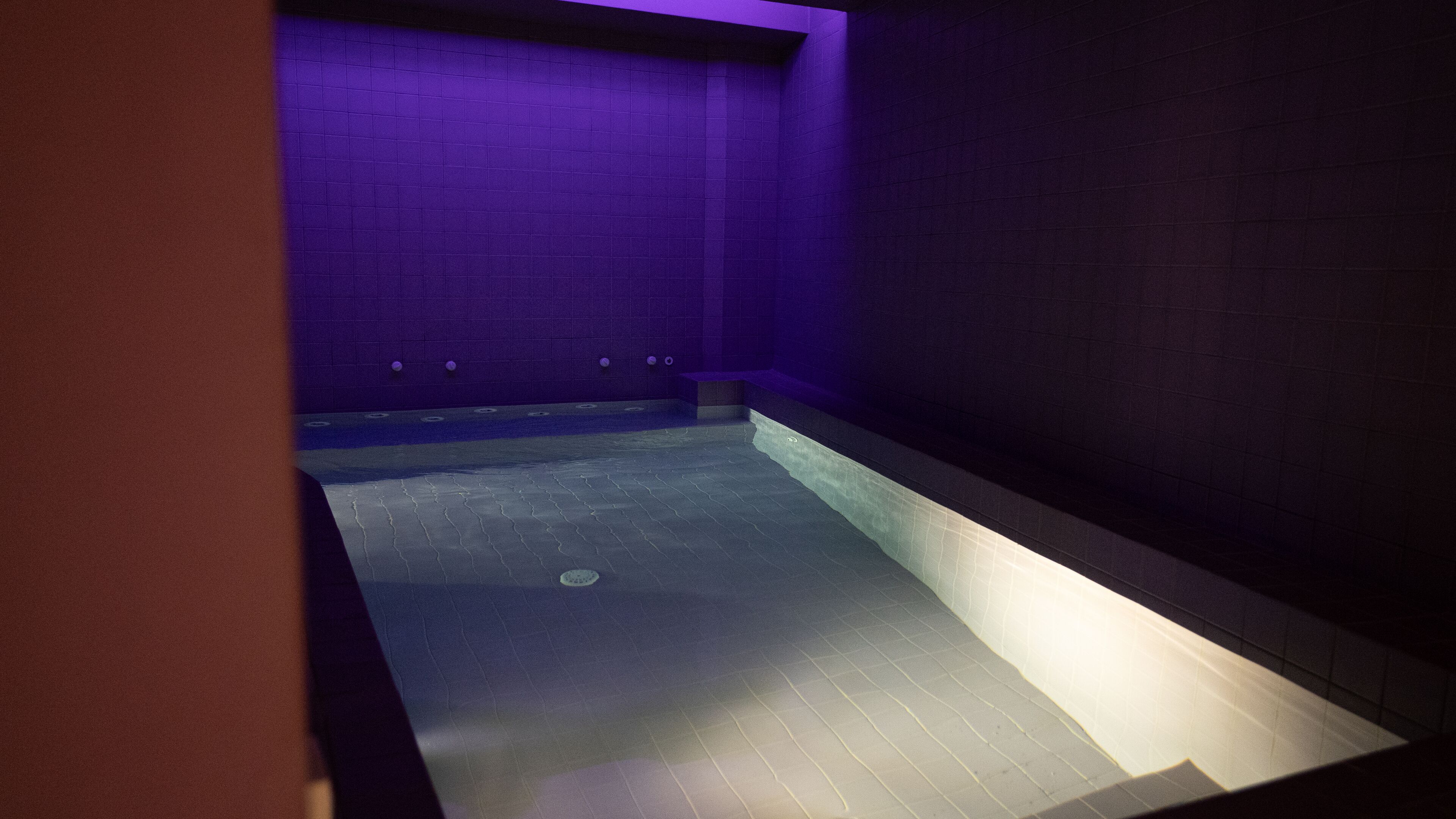 Un espace intérieur abritant une piscine, faiblement éclairé, avec des murs carrelés et un sol en pente menant à une petite piscine.
