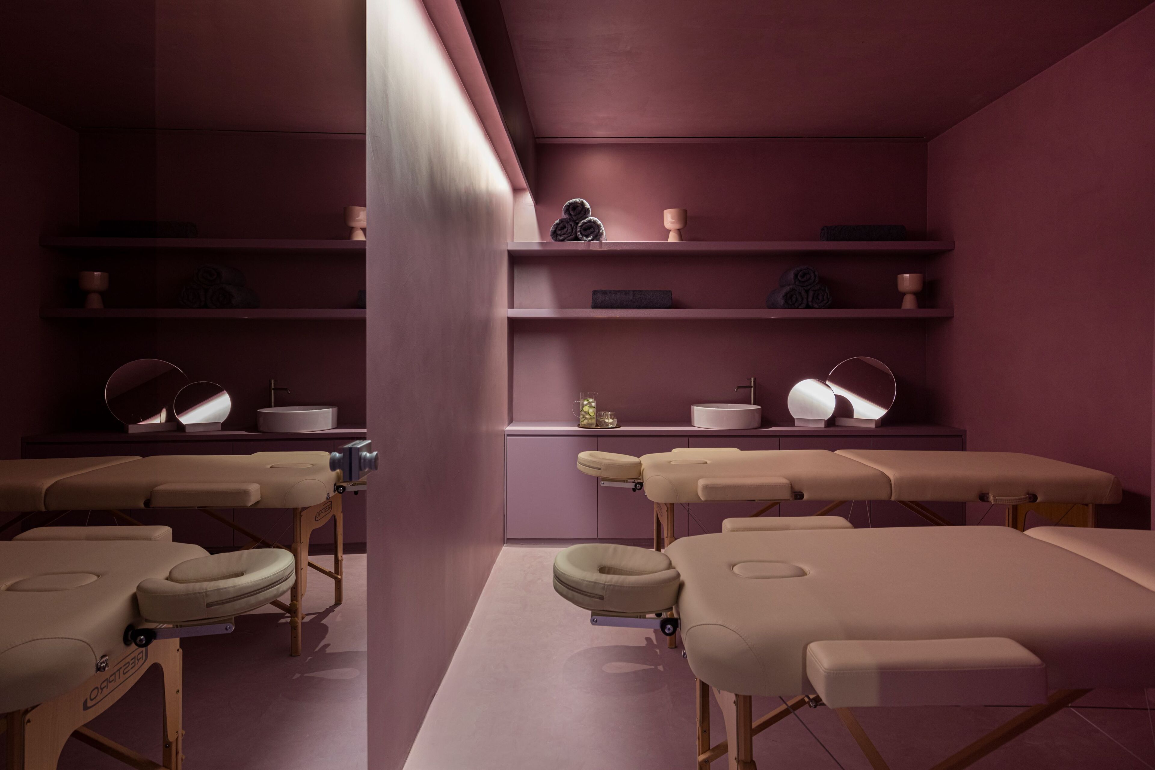 A clinical examination room with multiple examination tables, shelves with medical supplies, and a single patient bed.