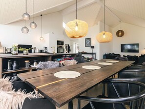 A modern kitchen with a dining area, featuring a long wooden table, hanging pendant lights, and a television mounted on the wall. - 5 Star Holiday Home in Stroby (Strøby)