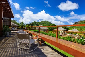 Une terrasse en bois avec des meubles d’extérieur, une structure à toit de chaume et une végétation luxuriante sous un ciel bleu dégagé.