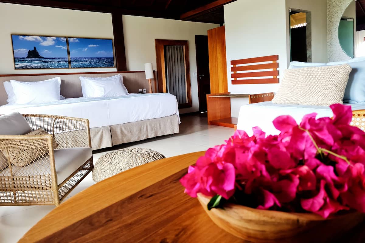 A hotel room with a bed, a chair, a table with flowers, and a view of the sea.