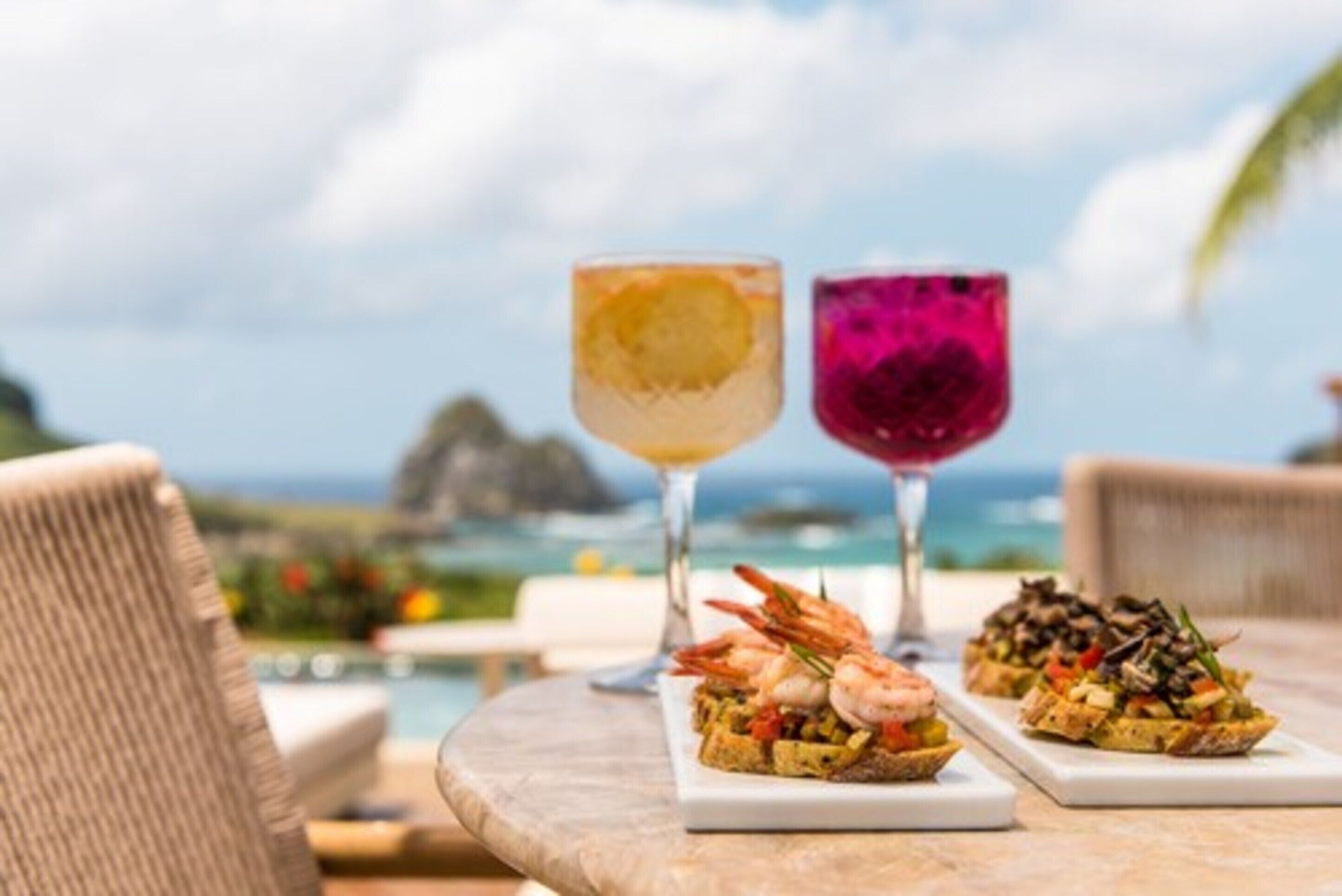 A table with two drinks and two dishes, set against a scenic ocean view.