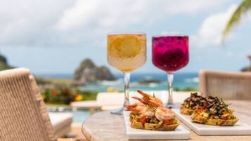 A table with two drinks and two dishes, set against a scenic ocean view.