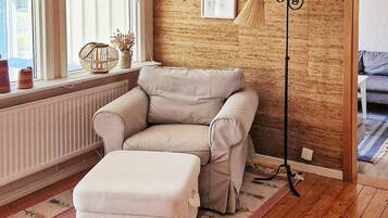 A room with a beige sofa, a matching ottoman, a floor lamp, a radiator, and a window with curtains.