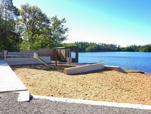A sandy beach area with a wooden structure, a swing set, and a small building by the water. - 6 Person Holiday Home in Trollhattan (Trollhattan)