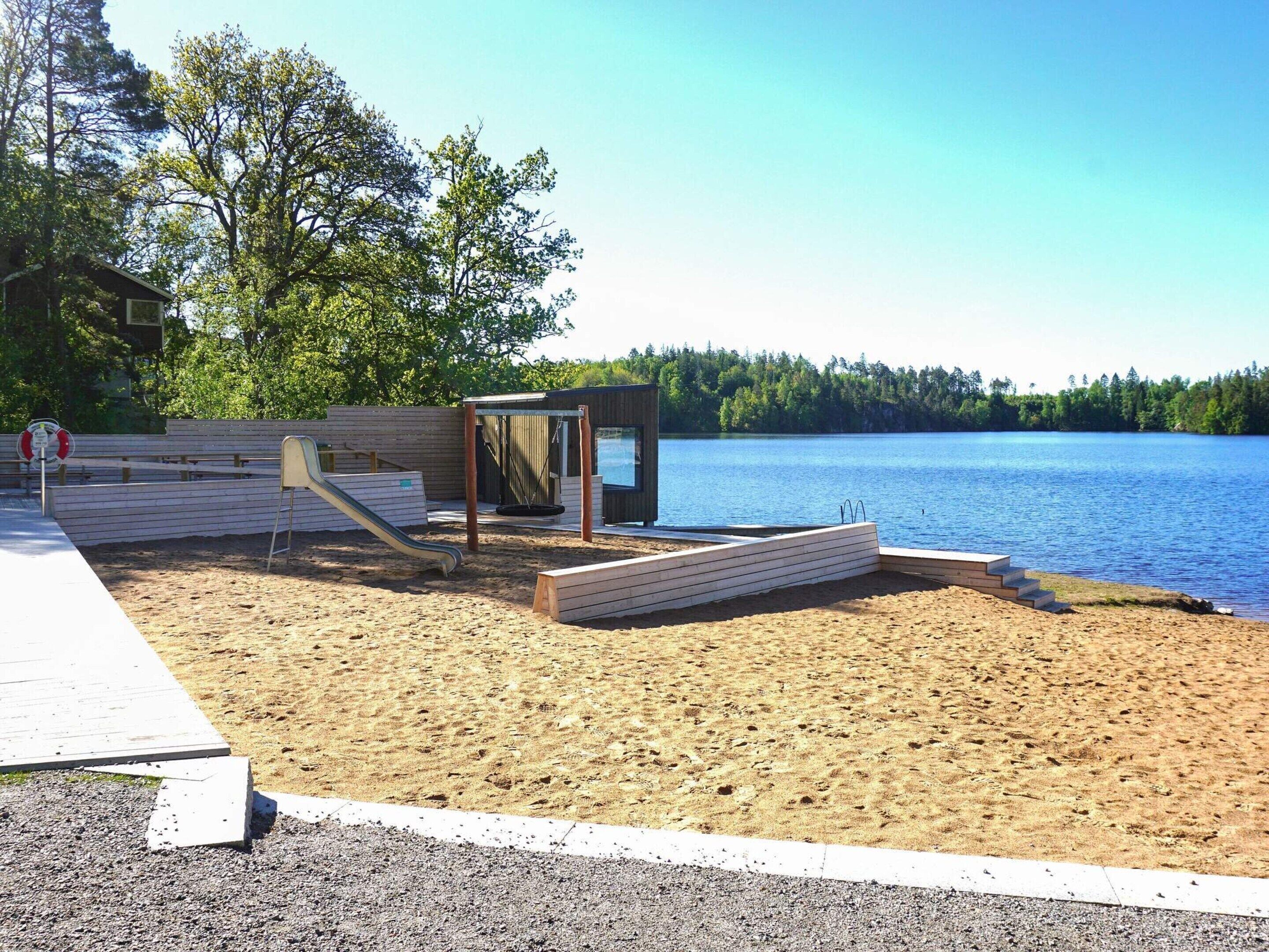 Zona de playa arenosa con una terraza de madera, un conjunto de columpios y una pequeña construcción de madera cerca del agua.