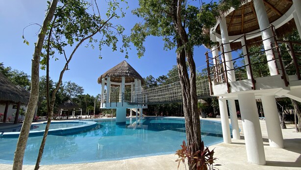 A resort pool area with a central water feature, surrounded by palm trees and a thatched-roof structure.