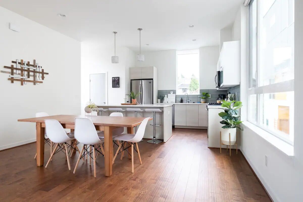 A modern kitchen with a wooden dining table and chairs, a stainless steel refrigerator, and a large window providing natural light.