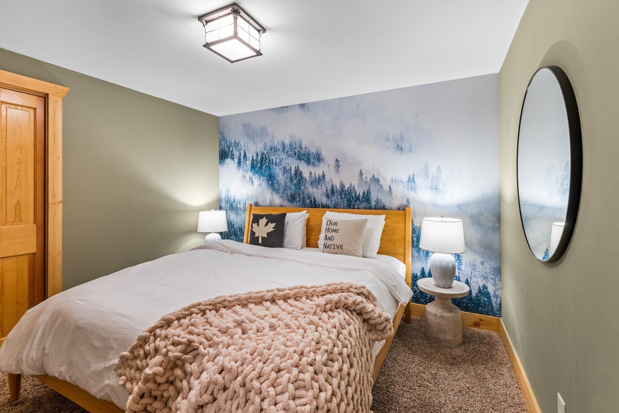 Een slaapkamer met een bed, een groot schilderij van een bos, een ronde spiegel en een houten garderobe.