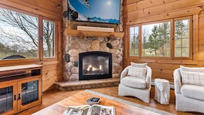 L’intérieur chaleureux d’un chalet avec une cheminée en pierre, un téléviseur fixé au mur et une vue sur l’extérieur grâce à de grandes fenêtres.