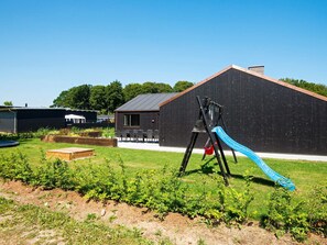 Une aire de jeux avec un toboggan et des balançoires, un bâtiment noir et une zone herbeuse avec des plantes.