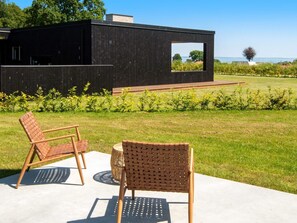 Een modern gebouw met een zwarte gevel, een klein raam en uitzicht op zee. Op de voorgrond staan twee rieten stoelen en een ronde tafel.