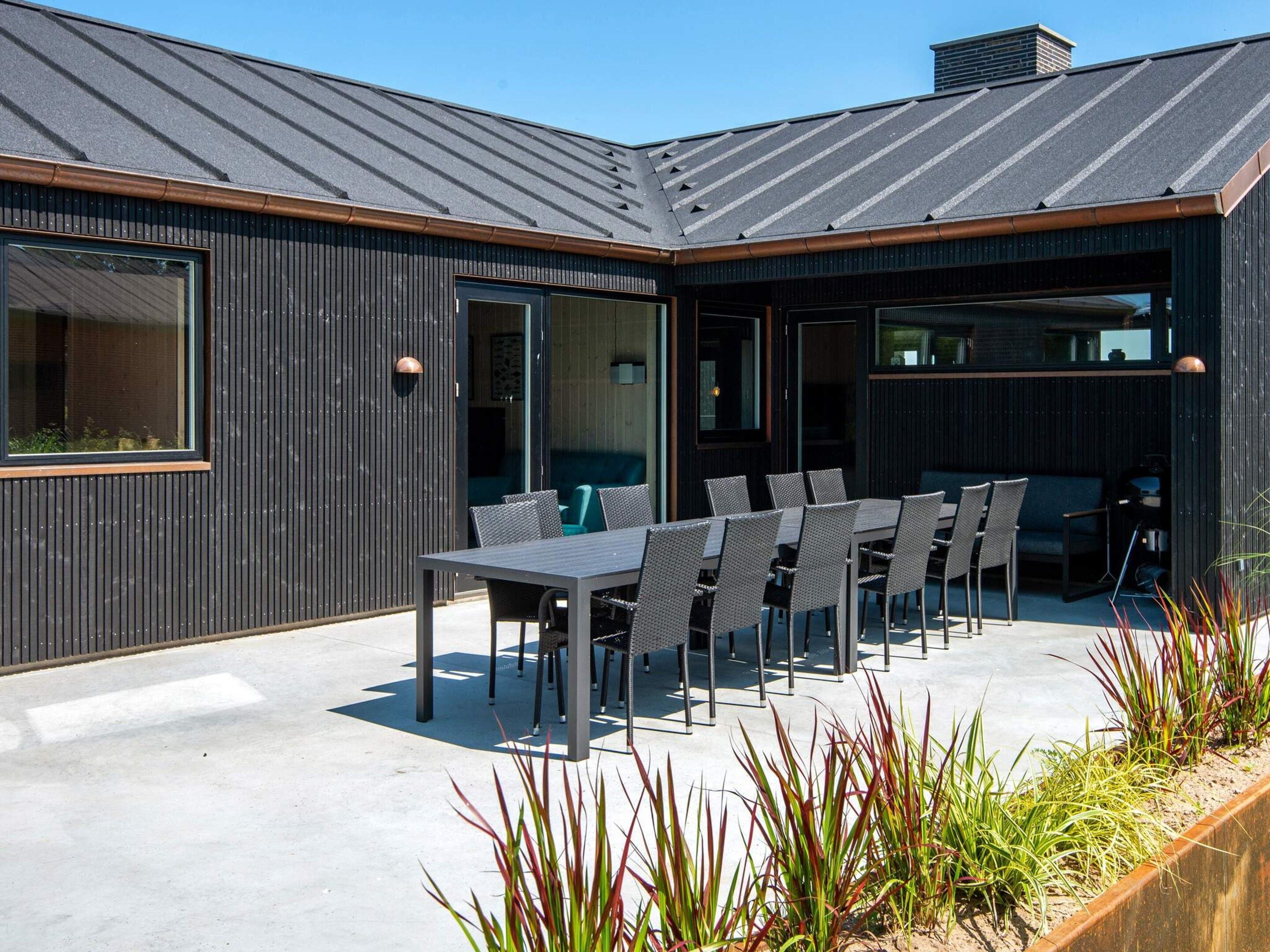 An outdoor dining area with a table and chairs, surrounded by a modern building with large windows and a metal roof.